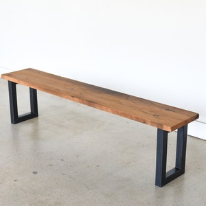 May include: A long wooden bench with a natural wood finish and black metal legs. The bench is made of reclaimed wood and has a rustic look.