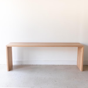 May include: A light-colored wooden console table with a rectangular top and two supporting legs. The table is made of natural wood and has a simple, modern design. The table is approximately 72 inches long.