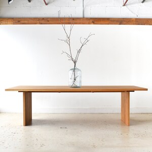 May include: A long, rectangular wooden table with a natural finish. The table has two legs on each end, and a vase with branches is centered on the table.