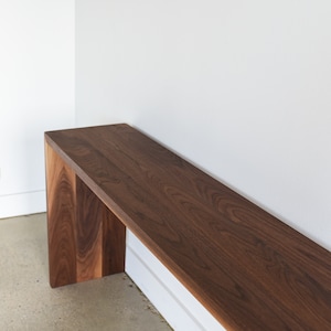 May include: A long, narrow wooden bench with a dark brown walnut finish. The bench has a simple, modern design with a rectangular top and two vertical supports.