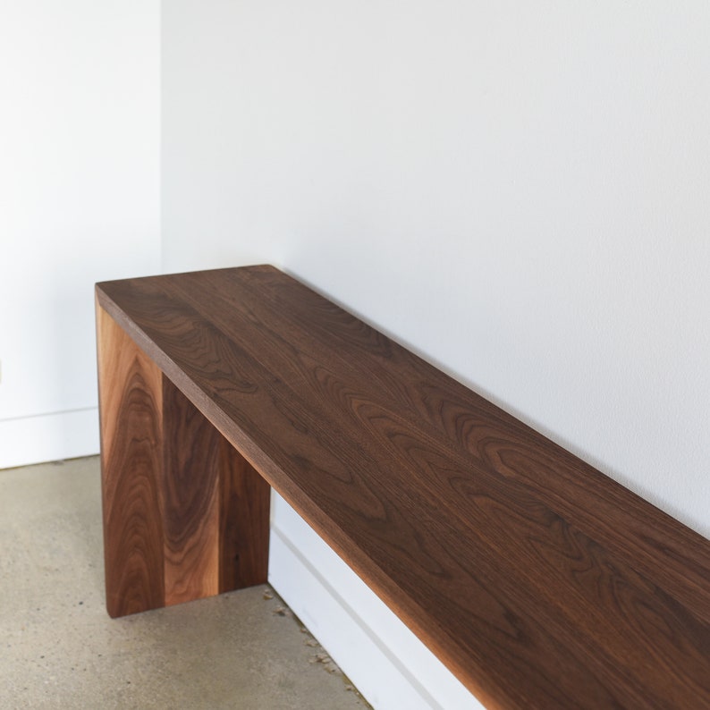 May include: A long, narrow wooden bench with a dark brown walnut finish. The bench has a simple, modern design with a rectangular top and two vertical supports.