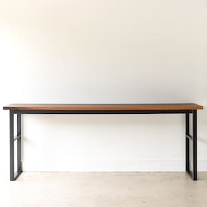 May include: A long, rectangular wooden table with a black metal frame. The table has a natural wood finish and is supported by two black metal legs on each end.