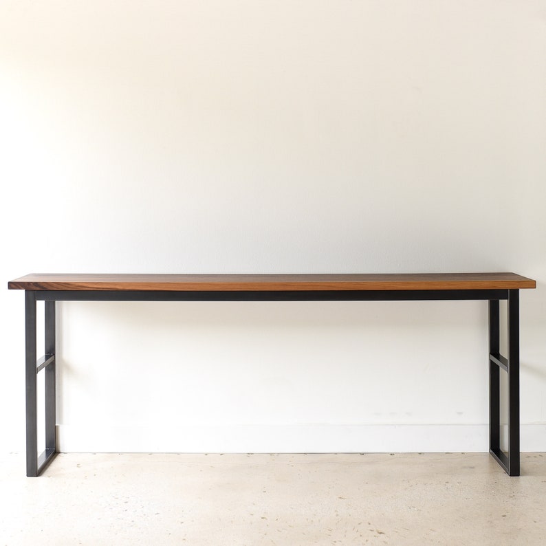 May include: A long, rectangular wooden table with a black metal frame. The table has a natural wood finish and is supported by two black metal legs on each end.