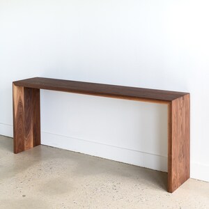 May include: A long, narrow console table made of dark walnut wood. The table has a simple, modern design with a rectangular top and a unique open frame base.