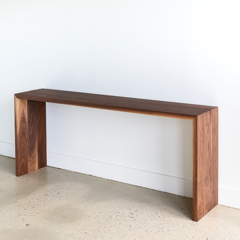 May include: A long, narrow console table made of dark walnut wood. The table has a simple, modern design with a rectangular top and a unique open frame base.