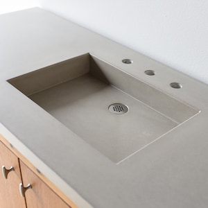 May include: A concrete bathroom sink with a rectangular basin and three holes for fixtures. The sink is set into a countertop made of wood.