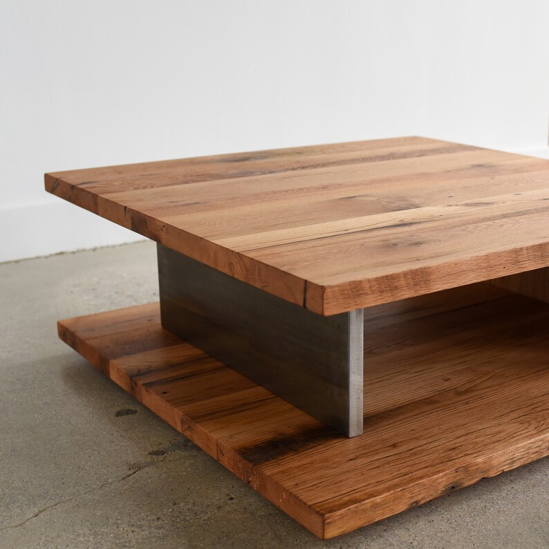 May include: A square coffee table featuring two thick, natural-toned wooden planks with visible grain patterns. The top and bottom wooden sections are separated by a central, vertical, dark gray metal support structure that connects the two levels.