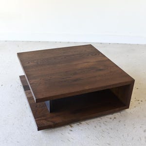May include: A square coffee table constructed from dark brown wood with a visible grain pattern. The table features a flat top surface and a lower shelf connected by a central support, creating an open, layered design with clean, straight edges.