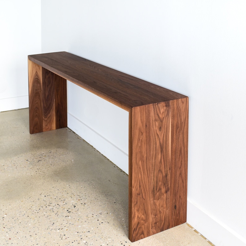 May include: A long, rectangular walnut wood console table with a simple, modern design. The table has a wide, flat top and two vertical supports on either end.