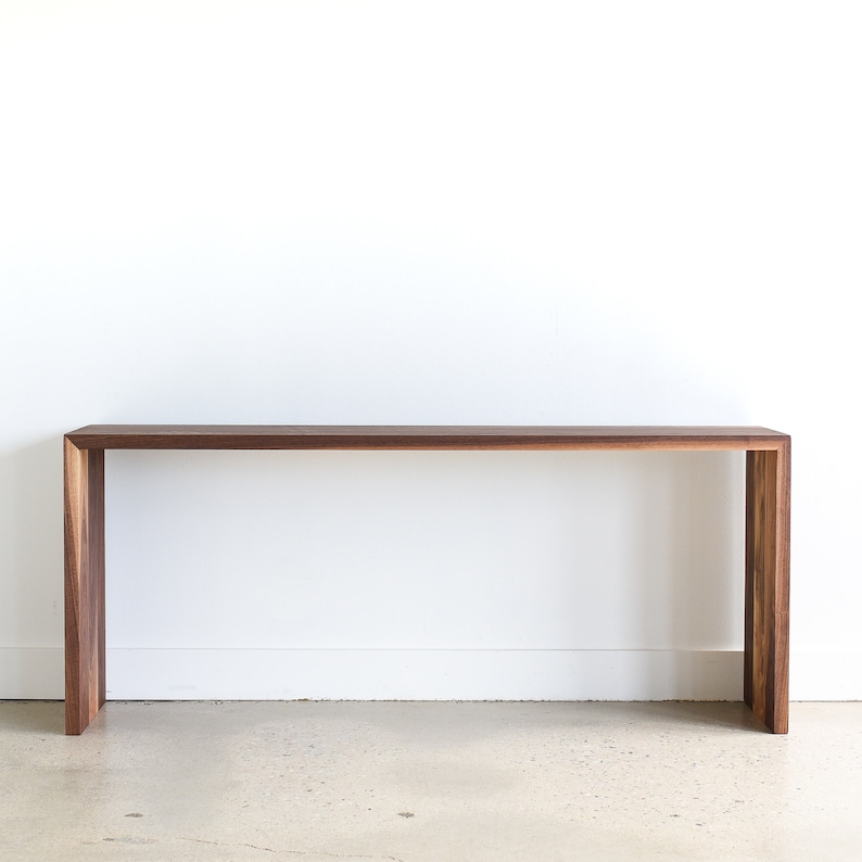 May include: A long, narrow console table made of dark walnut wood with a simple, modern design. The table has a rectangular top and two vertical supports at each end.