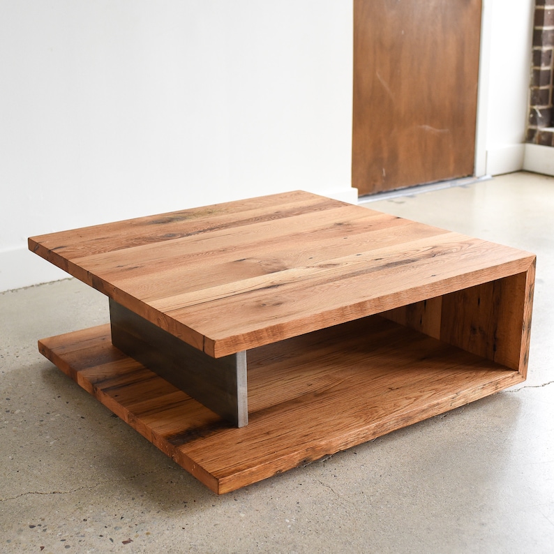 May include: A square coffee table constructed from thick, natural-toned wood planks featuring visible grain and knots. The table consists of a flat top and a lower base connected by a central, vertical dark gray metal support beam.