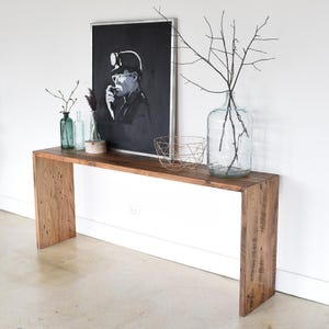 May include: A long, narrow wooden console table with a rustic finish. The table has a single shelf and is decorated with a black and white framed print, a clear glass vase with branches, a small metal bowl, and two glass bottles.