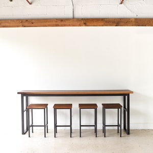 May include: A long, wooden table with a black metal frame and four matching wooden stools. The table and stools are in a white room with a wooden beam overhead.