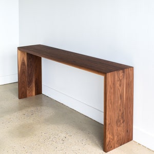 May include: A long, narrow console table made of dark walnut wood. The table has a simple, modern design with a rectangular top and a unique, open frame.