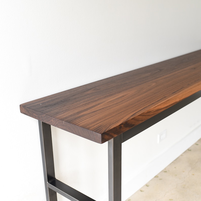 May include: A long, narrow console table with a dark wood top and a black metal frame. The table has a simple, modern design and would be a great addition to any home.