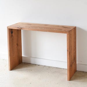 May include: A simple, modern console table made of solid wood with a natural finish. The table has a rectangular top and a rectangular base with open sides. The table is standing on a concrete floor.