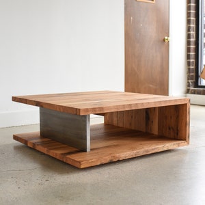 May include: A square coffee table constructed from thick, natural-toned wood planks featuring a visible grain. The table is supported by a central, vertical brushed metal plate that connects the top surface to the base, creating an open side design.