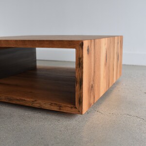 May include: A low, rectangular coffee table constructed from light brown wood with visible grain patterns and dark knots. The piece features a hollow, open-sided design with mitered corner joints, creating a continuous wood grain flow.
