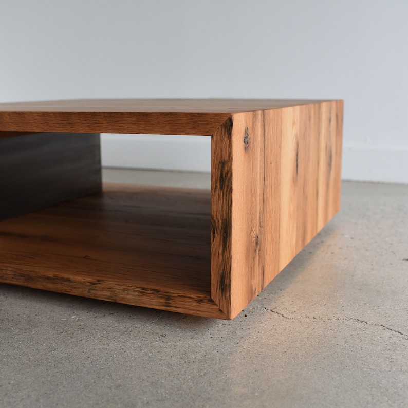 May include: A low, rectangular coffee table constructed from light brown wood with visible grain patterns and dark knots. The piece features a hollow, open-sided design with mitered corner joints, creating a continuous wood grain flow.
