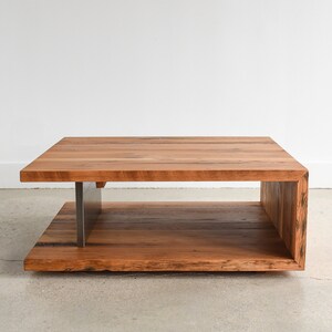 May include: A rectangular coffee table constructed from natural brown wood planks with a visible grain. The table features a C-shaped design with a solid wood top and base connected by a vertical brushed metal support on the left side.