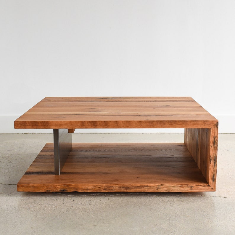May include: A rectangular coffee table constructed from natural brown wood planks with a visible grain. The table features a C-shaped design with a solid wood top and base connected by a vertical brushed metal support on the left side.