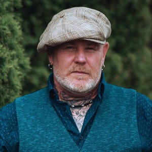 Pu&ograve; includere: Una persona con un berretto a quadri beige, un gilet verde acqua e una camicia blu scuro abbottonata. La persona ha barba, tatuaggi e una collana. Lo sfondo &egrave; un fogliame verde sfocato.