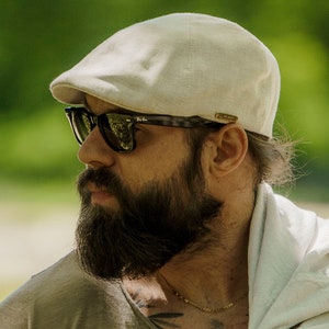 May include: A light beige newsboy cap and dark sunglasses. The person is wearing a green t-shirt and a white shirt draped over the shoulder. A watch with a black strap is visible on the wrist.