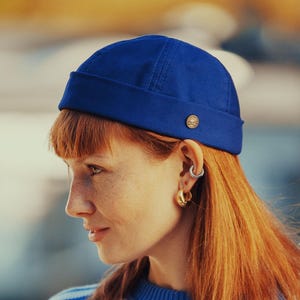 May include: A person with red hair wears a blue sailor hat with a gold button. They are also wearing a blue and white striped sweater with a nautical design.
