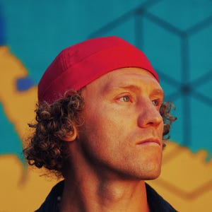 May include: A person wearing a red beanie, a navy blue corduroy shirt over a white t-shirt. The background features a map design with blue and yellow hues. The person is looking off to the side.