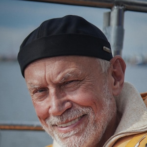 May include: A black docker cap featuring a small metallic side detail, worn with a bright yellow hooded jacket over a beige ribbed zip-up sweater. The cap has a structured, brimless design with a rolled edge, providing a clean and simple look.