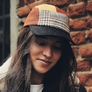 Puede incluir: Gorra de paneles múltiples con visera negra, parte superior marrón y amarilla y centro estampado. La gorra la lleva una persona con pelo largo castaño, sobre un fondo de pared de ladrillo. El diseño de la gorra incluye un estampado de pata de gallo.