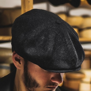 May include: Close-up view of a dark charcoal gray wool newsboy cap. The cap features a classic eight-panel design with a slightly curved brim.  The fabric appears to be soft and textured. The cap is shown against a blurred background of wooden shelves filled with various hats.