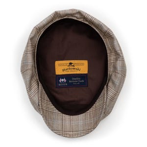 Pu&ograve; includere: Un cappello newsboy a quadri marroni e beige con interno marrone scuro. Il cappello ha una corona arrotondata e una visiera corta e rigida. L'etichetta interna recita "Sterkowski" e "Quality British Cloth".