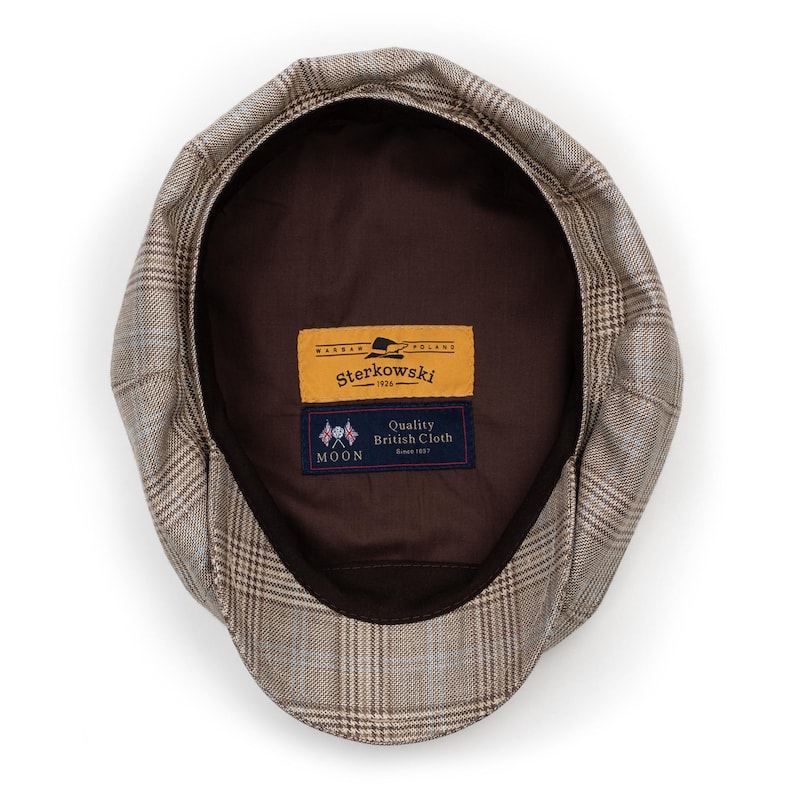 Pu&ograve; includere: Un cappello newsboy a quadri marroni e beige con interno marrone scuro. Il cappello ha una corona arrotondata e una visiera corta e rigida. L'etichetta interna recita "Sterkowski" e "Quality British Cloth".