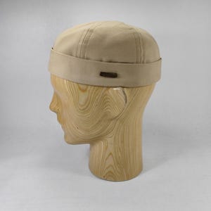 May include: A tan, unisex cap with a short brim and a small, rectangular brown accent. The cap is displayed on a wooden head form against a plain background. The cap has vertical stitching on the crown.