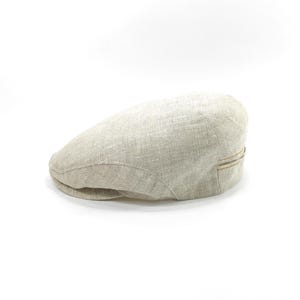 May include: A light beige newsboy cap made of a textured fabric. The cap has a rounded crown and a small, stiff brim. A thin, decorative band is sewn on the back of the cap. The cap is set against a white background.