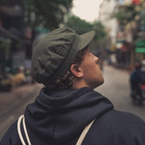 Op de afbeelding: Een groene pet met een korte klep. De pet wordt gedragen door een persoon die een marineblauwe hoodie draagt. De persoon kijkt omhoog, met een vage straatscène op de achtergrond.