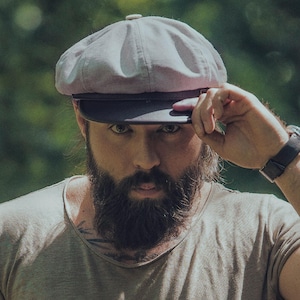 May include: A light gray newsboy cap with a dark brim. The cap is worn by a person with a full beard and a neutral-colored shirt. The background is blurred green foliage.