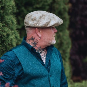 May include: A tan and brown plaid newsboy cap. The cap is worn by a person wearing a teal vest over a blue button-down shirt. The person has a beard, tattoos, and a silver hoop earring.