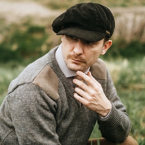 May include: A man wearing a brown corduroy newsboy cap and a grey sweater with a brown leather patch on the shoulder. He is looking down and has a hand on his chin.