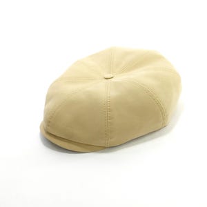 May include: A tan-colored, eight-panel newsboy cap with a small, curved brim. The cap features stitched seams radiating from a central button on the crown. The hat is made of a smooth, solid-colored fabric.