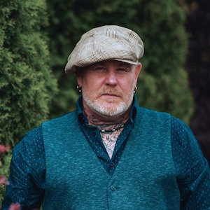 May include: A person wearing a tan plaid newsboy cap, teal vest, and a dark blue button-down shirt. The person has a beard, tattoos, and a necklace. The background is a blurred green foliage.