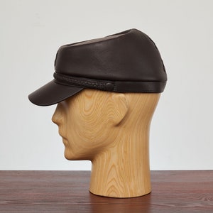 May include: A brown leather cap with a braided band around the crown. The cap is on a wooden mannequin head.