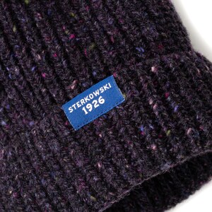 May include: Close-up of a dark purple knit beanie with flecks of color throughout. A blue rectangular tag with the text "STERKOWSKI 1926" is sewn on the front. The hat has a ribbed texture.