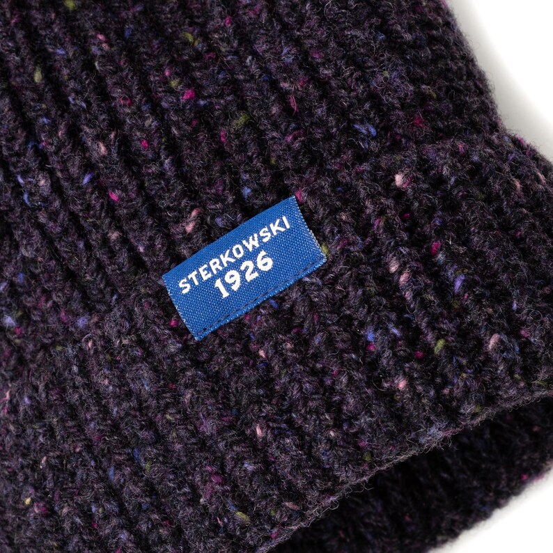 May include: Close-up of a dark purple knit beanie with flecks of color throughout. A blue rectangular tag with the text "STERKOWSKI 1926" is sewn on the front. The hat has a ribbed texture.
