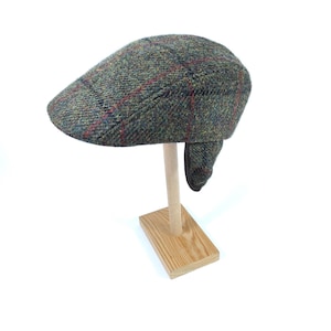 May include: A green tweed flat cap with a red and blue check pattern. The cap features ear flaps and is displayed on a wooden stand. A traditional style of hat, ideal for colder months.