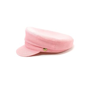 May include: A light pink, classic-style hat with a short brim and a rounded crown. The hat features a decorative band and a gold-colored button on the side. This hat is a stylish accessory.