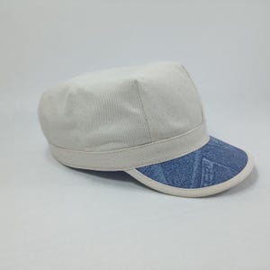 May include: A white canvas cap with a blue denim brim. The brim has a white trim around the edge.