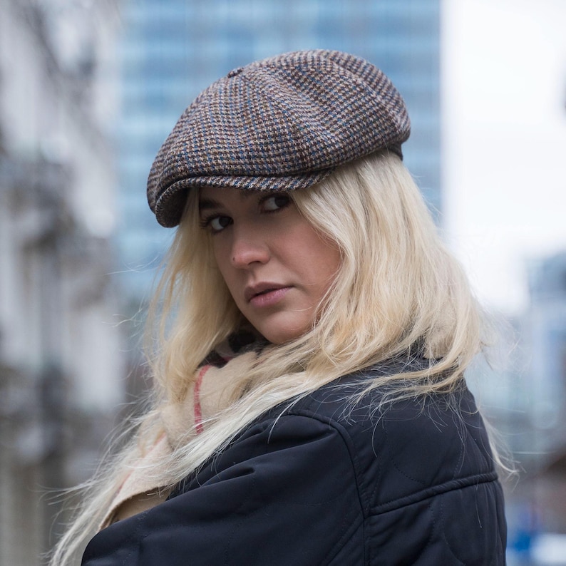 Puede incluir: Una mujer con cabello rubio lleva una gorra de newsboy de tweed marr&oacute;n y azul. Lleva una chaqueta negra y una bufanda con rayas rojas y blancas.