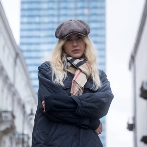 Puede incluir: Una mujer con una gorra de conductor marr&oacute;n, una chaqueta negra y una bufanda de cuadros crema y roja. Est&aacute; de pie frente a un edificio alto.